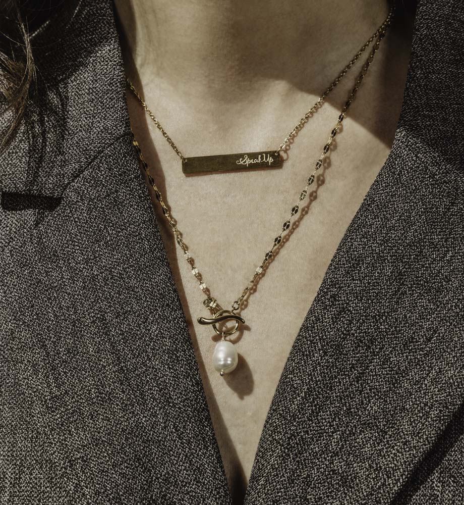 woman wearing gray blazer with two gold necklaces with pearls