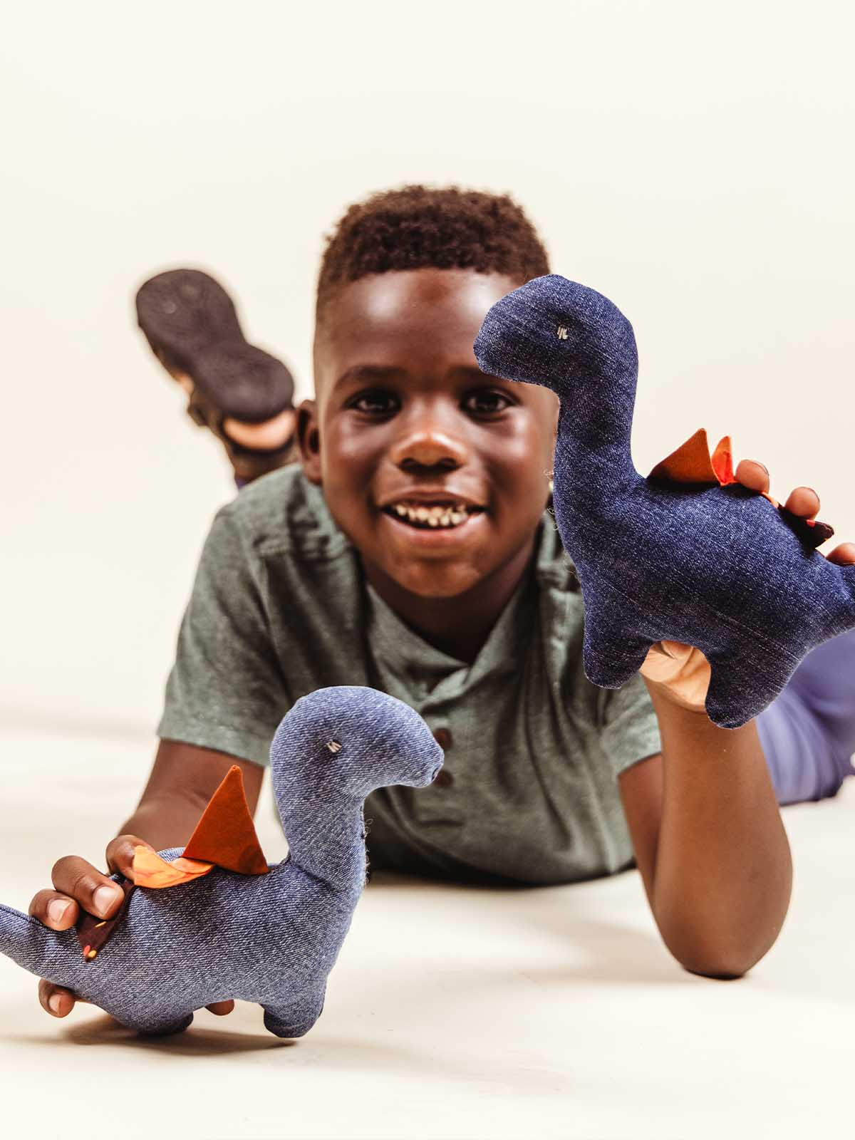 A smiling boy lies on his stomach holding up two blue fabric dinosaur plush toys with patterned spikes.