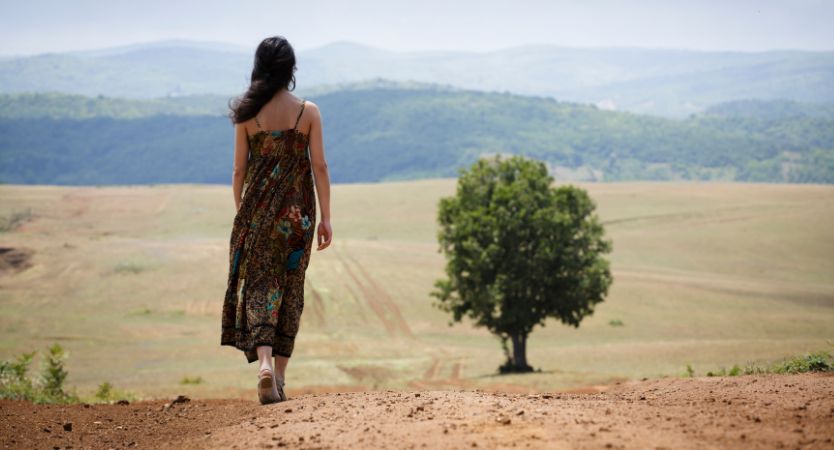 woman walking away down a dirty road with a tree on the right side of the image frame. 