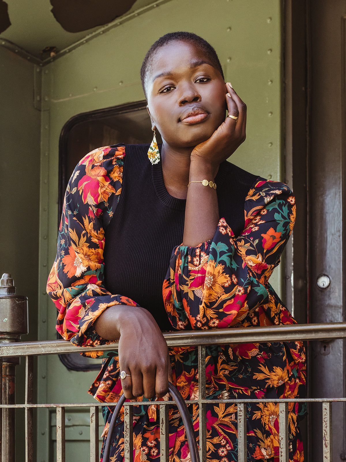 A woman with short hair leans against a metal railing, wearing a black top with a colorful floral-patterned blouse over it. She rests her face on her hand, showing off gold jewelry, including a bracelet, ring, and vibrant beaded earrings. Her expression is serene, and the background includes a vintage green train car, adding a nostalgic feel to the scene.