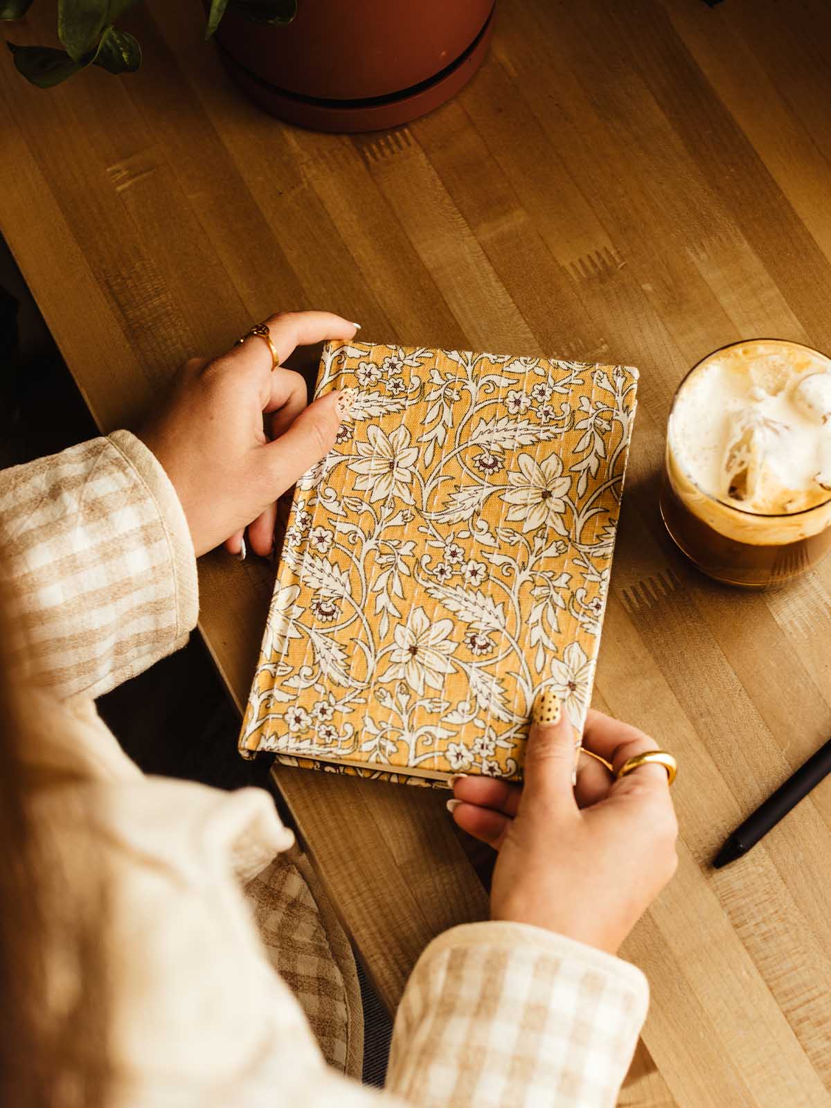 A person holding a hardcover journal with a mustard yellow and beige floral pattern, set on a wooden table next to a cup of iced coffee.