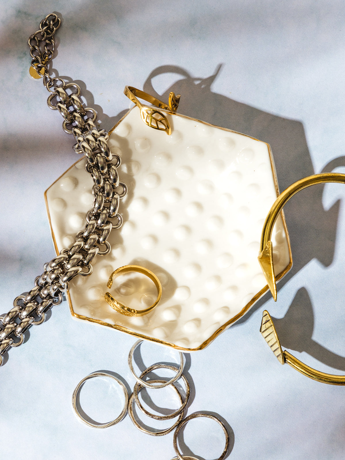 Gold-trimmed ceramic jewelry dish with textured surface, styled with mixed metal rings, bracelets, and a gold leaf cuff on a blue backdrop.